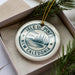 A white ornament with a blue design, featuring the text "ISLE OF PINES NEW CALEDONIA" and a landscape with pine trees and a body of water.
