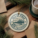 A circular ornament with the text "ISLE OF PINES NEW CALEDONIA" printed on it, surrounded by pine branches and a cup of coffee on a wooden surface.