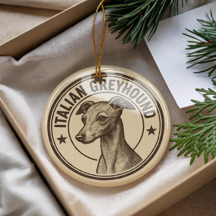 A circular ornament featuring an illustration of a greyhound dog, with the text "Italian Greyhound" printed on it, placed inside a box with pine branches.