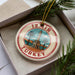 A circular ornament with a red border and a picture of a tower, surrounded by palm trees, is displayed in a box with pine branches.
