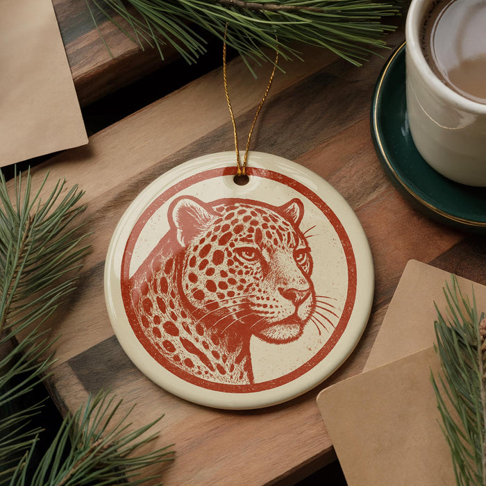 a round ornament with a leopard's face on it, placed on a wooden surface next to a cup of coffee and some pine branches.