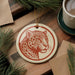 a round ornament with a leopard's face on it, placed on a wooden surface next to a cup of coffee and some pine branches.