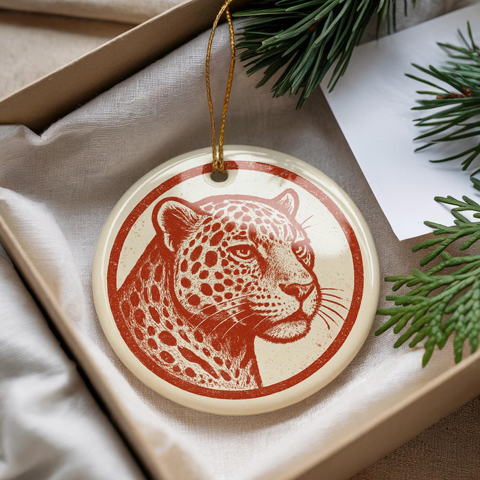 A round ornament featuring a leopard's face is displayed in a box, surrounded by pine branches.