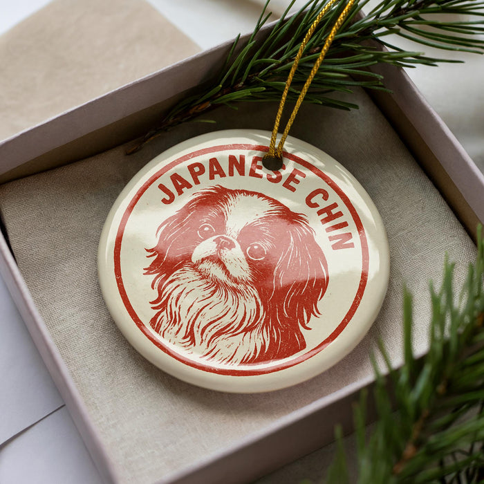 A circular ornament with a dog's face and the text "JAPANESE CHIN" on it, placed inside a box with pine branches.