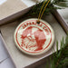 A circular ornament with a dog's face and the text "JAPANESE CHIN" on it, placed inside a box with pine branches.