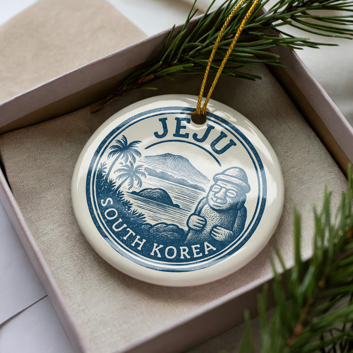 A decorative ornament in a box, featuring a circular design with a blue border and a white background. The design includes a depiction of a mountain, palm trees, and a figure wearing a hat. The text "JEJU SOUTH KOREA" is prominently displayed on the ornament.