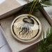 A round ornament with a black and white illustration of a jellyfish is displayed in a box, surrounded by pine branches.