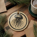 a round ornament with a black and white illustration of a jellyfish, hanging on a wooden surface. There is also a cup of coffee and some pine branches in the background.