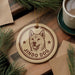 A round ornament featuring a dog's face and the text "JINDO DOG" is displayed on a wooden surface, surrounded by pine branches and a cup of coffee.