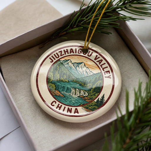 A decorative ornament in a box, featuring a scenic landscape with mountains, waterfalls, and pine trees.