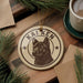 A circular ornament featuring a black and white image of a dog's head, with the name "KAIKEN" written around the edge. The ornament is hanging from a string, and there is a cup of coffee and some pine branches in the background.