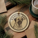 A circular ornament featuring a kangaroo dog illustration, placed on a wooden surface with pine branches and a cup of coffee nearby.