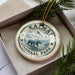 A circular ornament with the text "KAIMAI NATIONAL PARK USA" printed on it, featuring a bear illustration. The ornament is placed inside a box with pine branches.