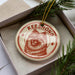 A circular ornament with a red and white design featuring a dog's face, placed inside a box with pine branches.