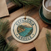 A decorative ornament with the text "Kefalonia Greece" and a scenic illustration of a coastal town, surrounded by pine branches and a cup of coffee.
