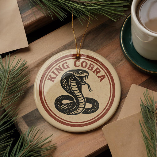 A round ornament with the text "KING cobra" and a graphic of a cobra is displayed on a wooden surface, surrounded by pine branches and a cup of coffee.