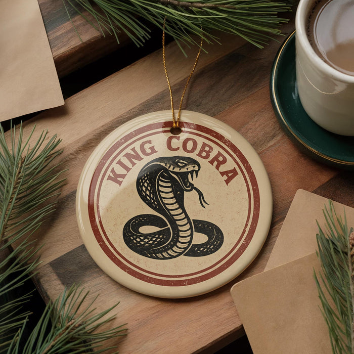 A round ornament with the text "KING cobra" and a graphic of a cobra is displayed on a wooden surface, surrounded by pine branches and a cup of coffee.