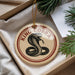 A round ornament with a black and white snake design, featuring the text "KING cobra" on it, is displayed in a box with pine branches.