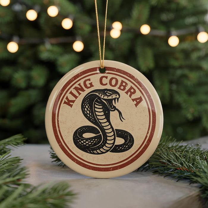 A round ornament with a red and white border, featuring a black and white illustration of a coiled snake, is displayed in front of a blurred Christmas tree.