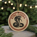 A round ornament with a red and white border, featuring a black and white illustration of a coiled snake, is displayed in front of a blurred Christmas tree.