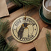 A round ornament featuring a penguin is displayed on a wooden surface, surrounded by pine branches and a cup of coffee.