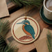 a round ornament with a bird design, placed on a wooden surface next to a cup of coffee and some pine branches.