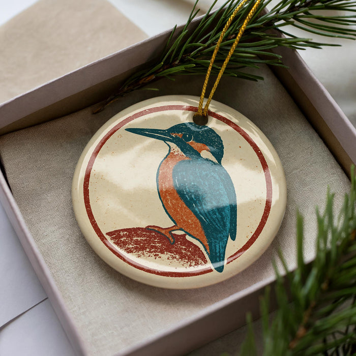 A decorative ornament featuring a colorful bird, likely a kingfisher, is displayed in a box surrounded by pine branches.