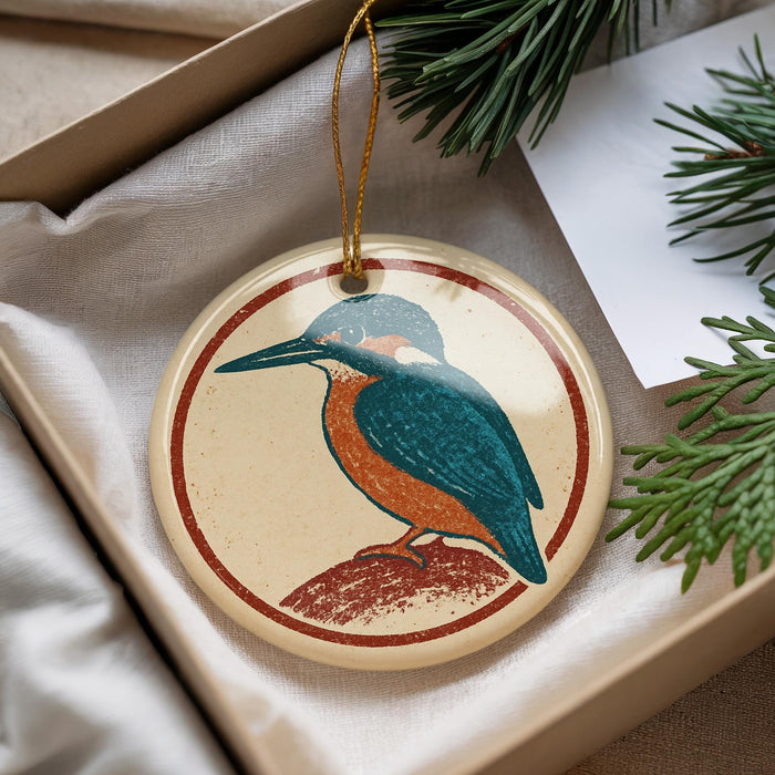 A round ornament featuring a colorful bird perched on a branch, placed in a box with pine branches and a white card.