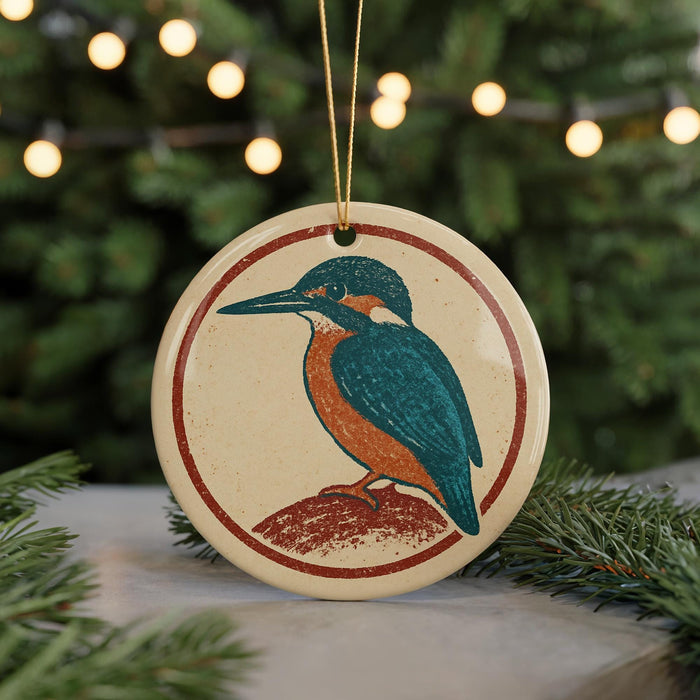 A round ornament featuring a colorful bird, likely a kingfisher, is displayed in front of a blurred Christmas tree with twinkling lights.