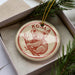 A circular ornament featuring a koala bear is displayed in a box, surrounded by pine branches.