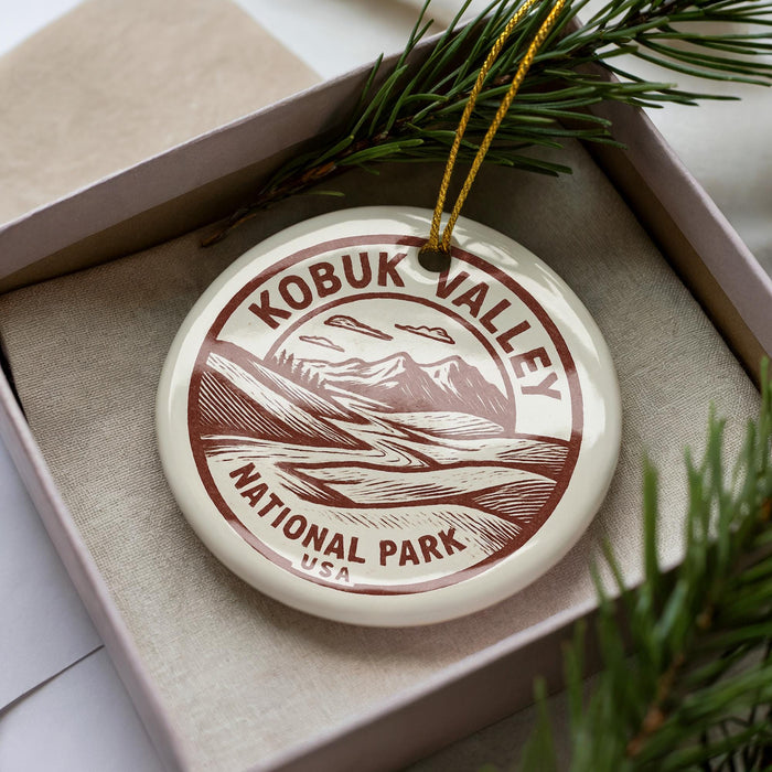 A circular ornament with the text "KOBUK VALLEY NATIONAL PARK USA" printed on it, placed inside a box with pine branches.