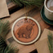 A circular ornament featuring a brown dog, possibly a Komondor, is displayed on a wooden surface alongside a cup of coffee and some pine branches.