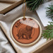 A circular ornament with a brown border and a picture of a shaggy dog, likely a Komondor, is displayed in a box with pine branches.