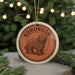 A circular ornament featuring a brown dog, possibly a Komondor, with the word "KOMONDO" written on it, hanging in front of a Christmas tree with twinkling lights.