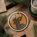 a decorative ornament with a deer or antelope design, hanging on a wooden surface. The ornament is circular in shape and has a gold string attached to it. There is also a cup of coffee and some pine branches in the background.