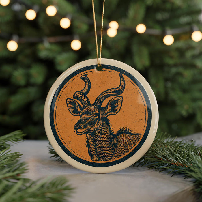 A ceramic ornament featuring a detailed illustration of a deer or antelope with large curved horns, hanging on a string and surrounded by pine branches.