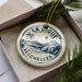 A circular ornament with a palm tree and mountains, featuring the text "LA DIGUE SEYCHELLES" on it, is displayed in a box with pine branches.
