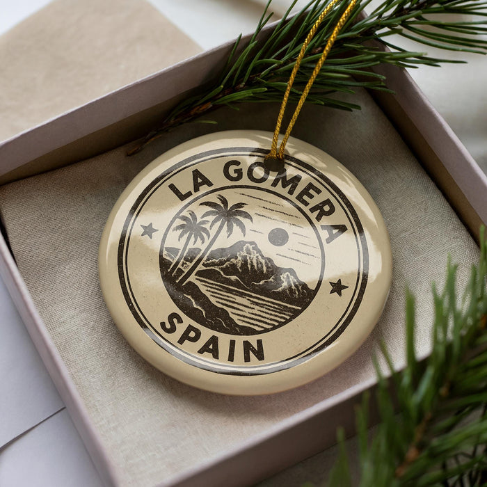 A circular ornament with the text "LA GOMERA SPAIN" printed on it, surrounded by palm trees and mountains, is displayed in a box with pine branches.