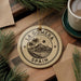 A circular ornament with the text "LA GOMERA SPAIN" printed on it, surrounded by pine branches and a cup of coffee on a wooden surface.