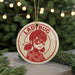 A red and white ornament featuring a cartoon image of a young girl with a mask, surrounded by Christmas lights and greenery.
