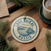 A circular ornament with the text "LAKE BAIKAL RUSSIA" printed on it, surrounded by pine branches and a cup of coffee on a wooden surface.