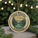 A circular ornament with a mountain landscape and a lake, surrounded by greenery, is displayed in front of a blurred background with Christmas lights.
