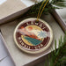 A circular ornament with a tropical island design, featuring palm trees, mountains, and a sunset, is displayed in a box with pine branches.