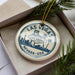 A circular ornament with the text "LAS VEGAS" and "WELCOME LAS VEGAS NEVADA, USA" printed on it, placed in a box with pine branches.