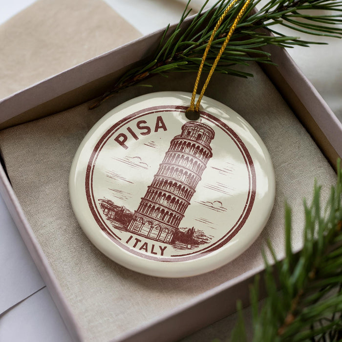 A circular ornament featuring the Leaning Tower of Pisa, a famous landmark in Italy, is displayed in a box with pine branches.