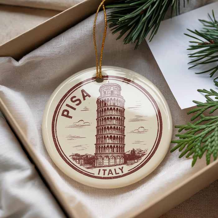 A circular ornament with the image of the Leaning Tower of Pisa, a famous landmark in Italy, is displayed in a box with pine branches.