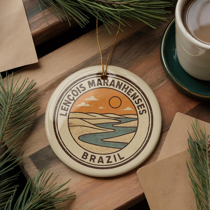 a circular ornament with a landscape design, including a river and mountains, and the text "LENCOIS MARANHENSES BRAZIL" around the edge. The ornament is placed on a wooden surface, with pine branches and a cup of coffee nearby.