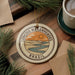 a circular ornament with a landscape design, including a river and mountains, and the text "LENCOIS MARANHENSES BRAZIL" around the edge. The ornament is placed on a wooden surface, with pine branches and a cup of coffee nearby.