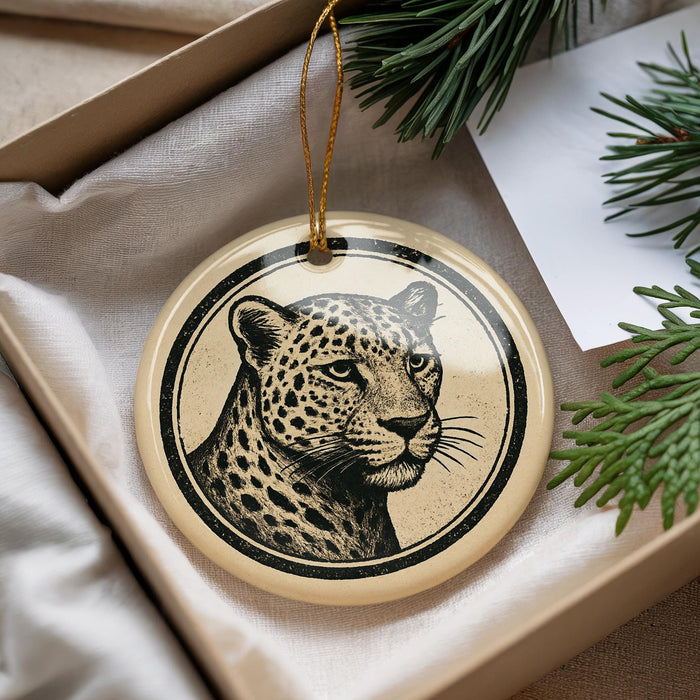 A round ornament featuring a leopard's face is displayed in a box, surrounded by pine branches.