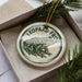 A leopard frog ornament is displayed in a box, surrounded by pine branches.